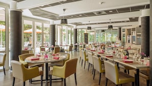 A spacious restaurant with round tables and yellow chairs, large windows, and a modern ceiling design.