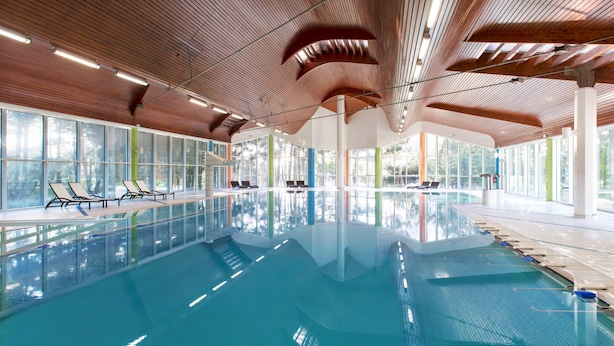 A large indoor swimming pool with wooden ceiling beams, clear glass walls, and lounge chairs along the poolside.