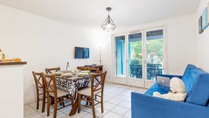 A dining area with a table set for four, a blue sofa with a teddy bear, and a view of the outdoors through a sliding glass door. - Duplex 8p • Réserve naturelle | Piscine incluse (Le Teich)