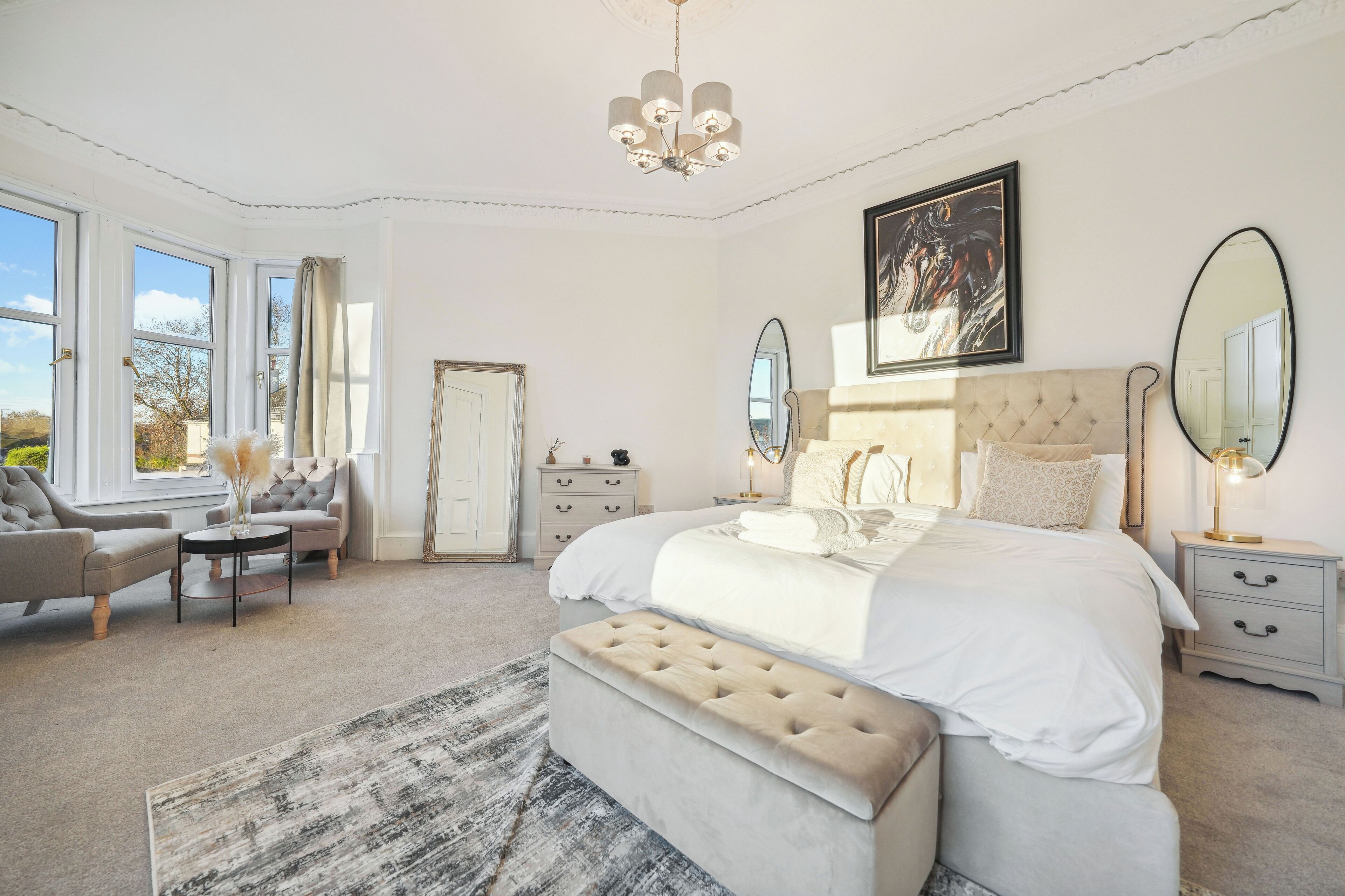 A bedroom with a large bed, a sitting area with chairs, a dresser, and a large window with curtains.