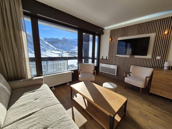 A modern living room with a sofa, armchairs, a coffee table, and a TV mounted on the wall. Large windows offer a view of a snowy mountain landscape.