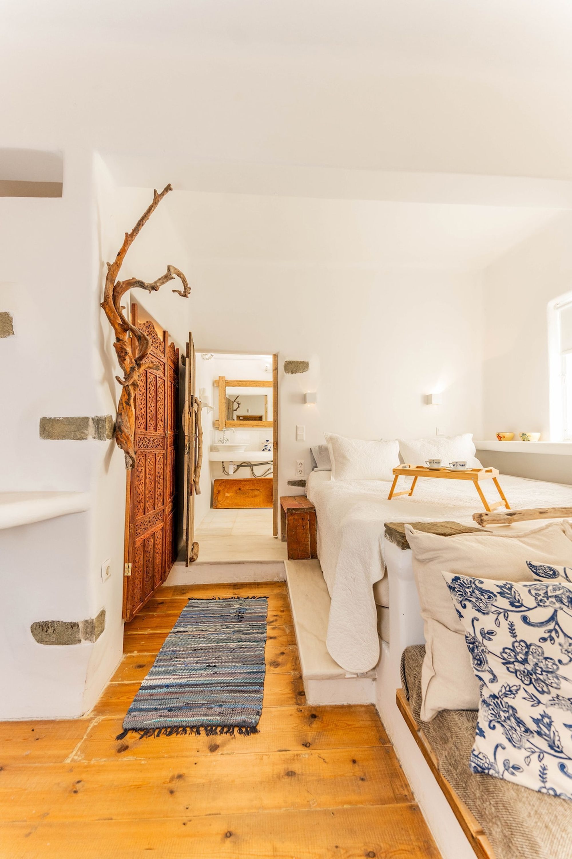 A cozy bedroom with a wooden floor, a bed with white bedding, a blue and white patterned pillow, a wooden tray on the bed, and a rustic door.