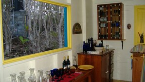 Ein Zimmer mit großem Fenster und Blick auf einen Wald, ein Holzkabinett mit Gläsern und ein Tisch mit Flaschen und Gläsern.