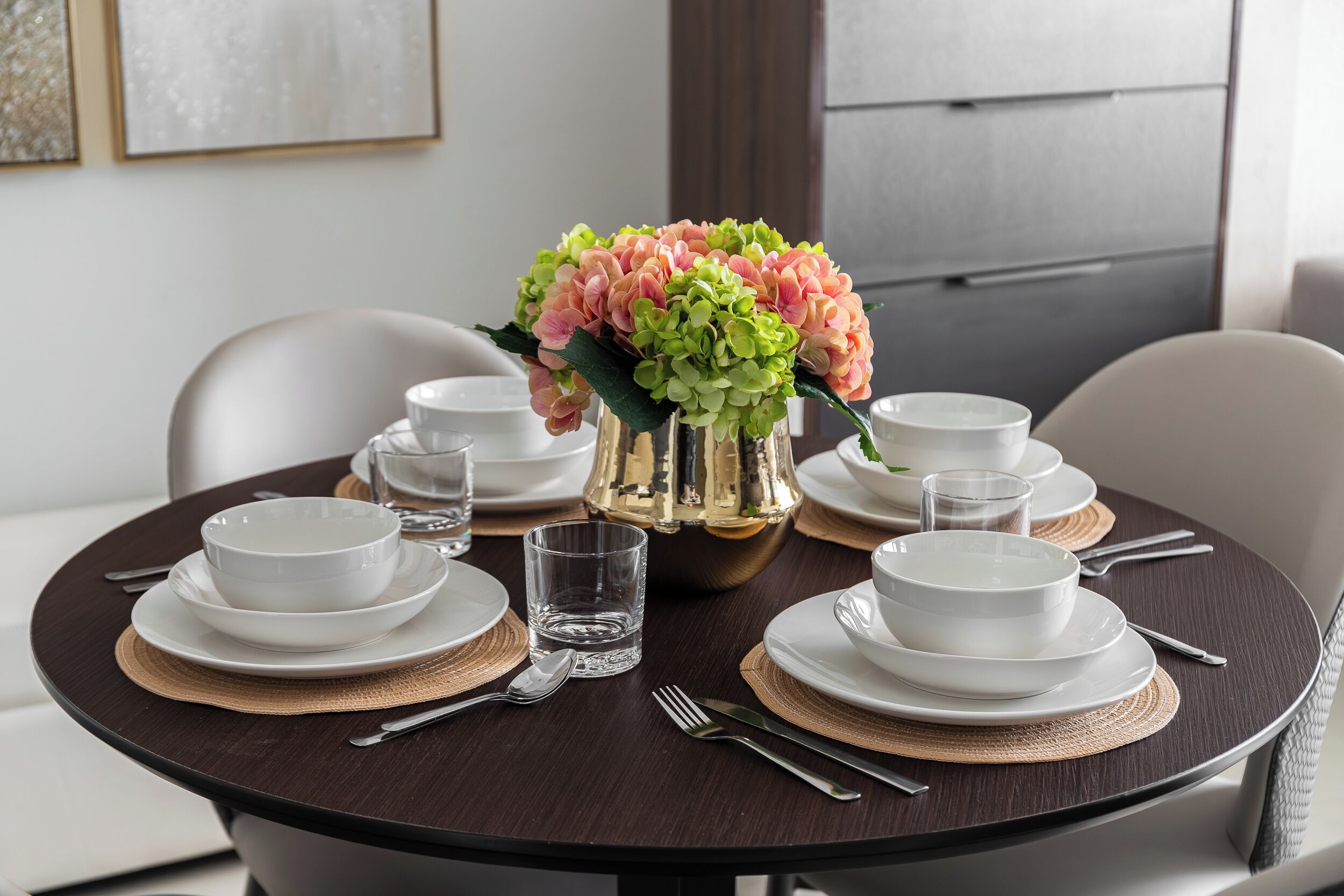 Een ronde eettafel met witte borden, kommen en bestek, een glas water en een vaas met roze en groene bloemen.