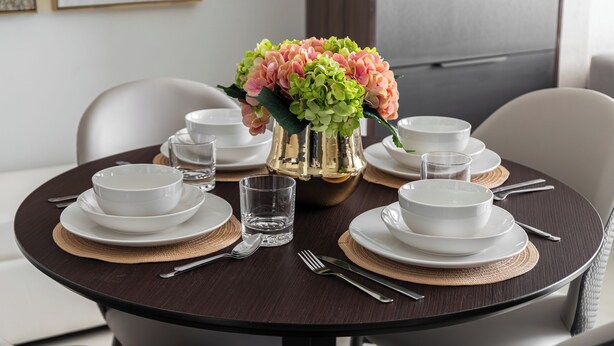 A round dining table with white plates, bowls, and cutlery, a glass of water, and a vase with pink and green flowers.