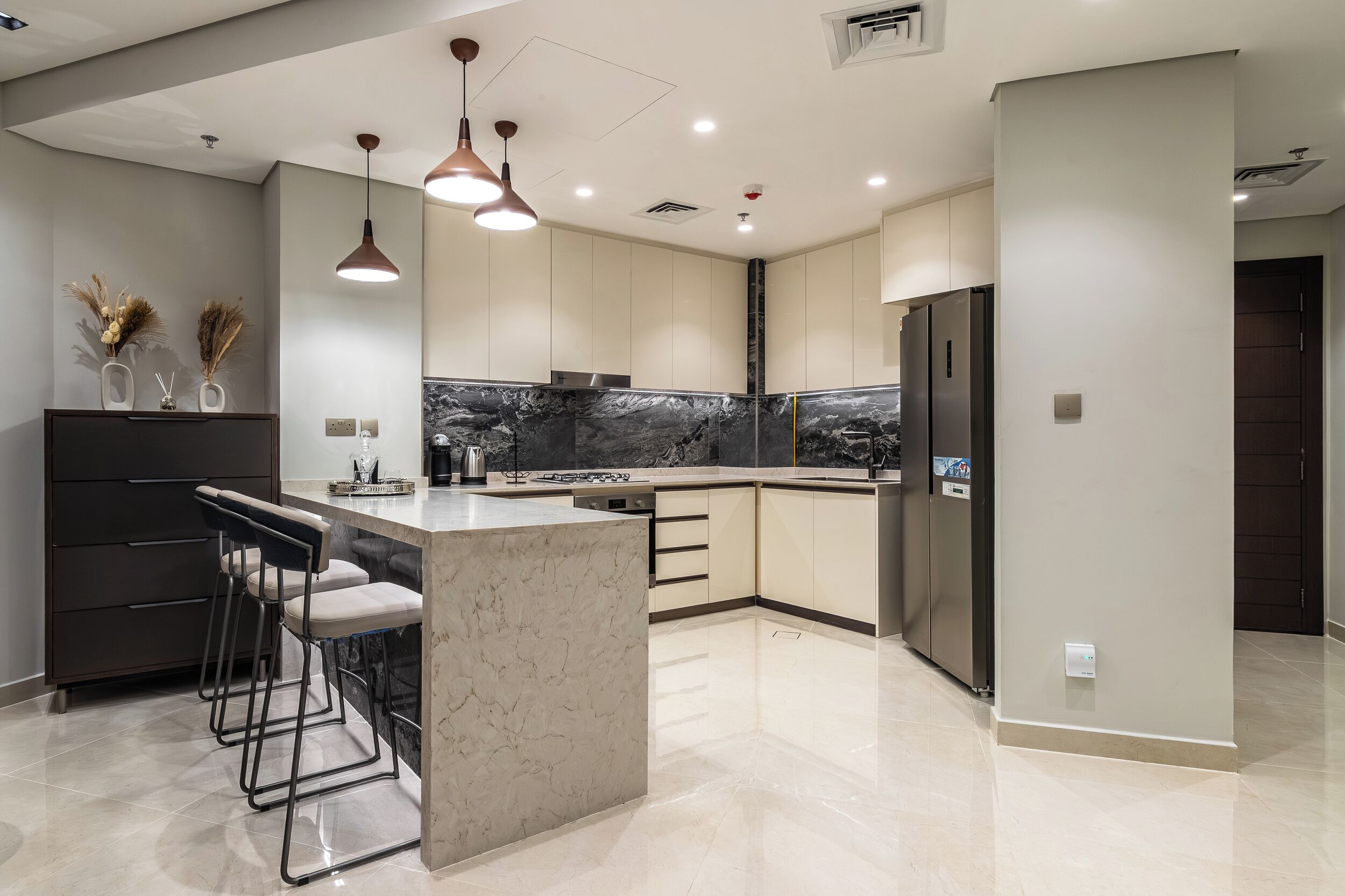 A modern kitchen with a marble island, bar stools, pendant lighting, and a built-in oven.