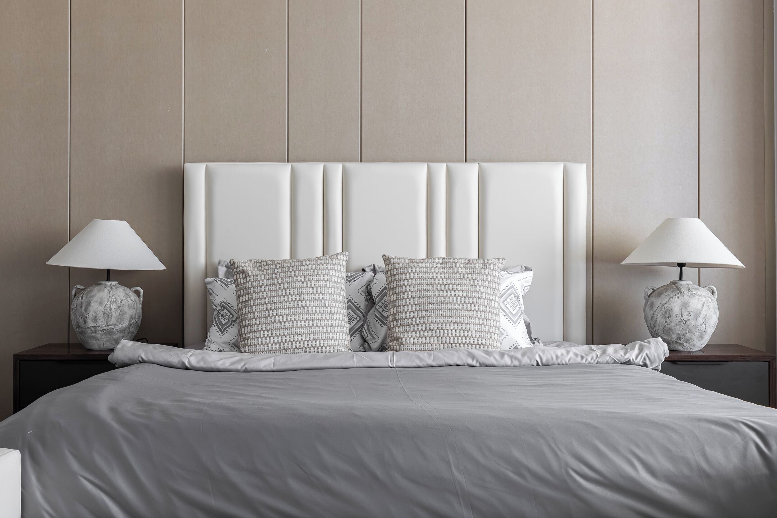 Una cama bien hecha con una cabecera acolchada, dos mesitas de noche con lámparas y almohadas estampadas.