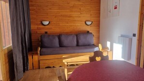 A cozy room with a wooden wall, a sofa with cushions, a wooden table, and a bed with a red cover. - Ski-in/ski-out & foot of the slopes, 5th floor, view ski slopes, balcony, tv, 28m², La Plagne (La Plagne)