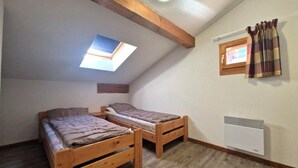 A small, cozy attic bedroom with two wooden beds, a skylight, a window with curtains, and a wall-mounted heater.