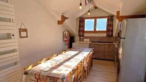 A dining area with a long table covered in a tablecloth, wooden chairs, a kitchen with a refrigerator, and a window with curtains. - 100m from ski slopes, 2nd floor, balcony, ski locker, 90m², Valfréjus (Valfréjus)
