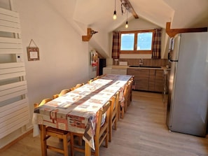 Ruokailutila, jossa on pitkä pöytä, jonka päällä on pöytäliina, puiset tuolit, keittiö, jossa on jääkaappi, ja ikkuna, jossa on verhot.