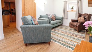 A living room with a sofa, armchair, and a patterned rug. There is a sliding door leading to a kitchen. - Bwthyn Taid (Holywell)
