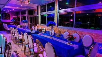 Una larga mesa con sillas, decorada con flores azules y un mantel blanco, en una habitación con ventanas grandes y una araña.