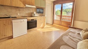 A compact kitchen with wooden cabinets, a dishwasher, and a stove. There's a television on the wall and a sliding glass door leading to a balcony with a view of greenery. - 140m from ski slopes, 2nd floor, view mountain, balcony, ski locker, 37m², Flumet (Flumet)
