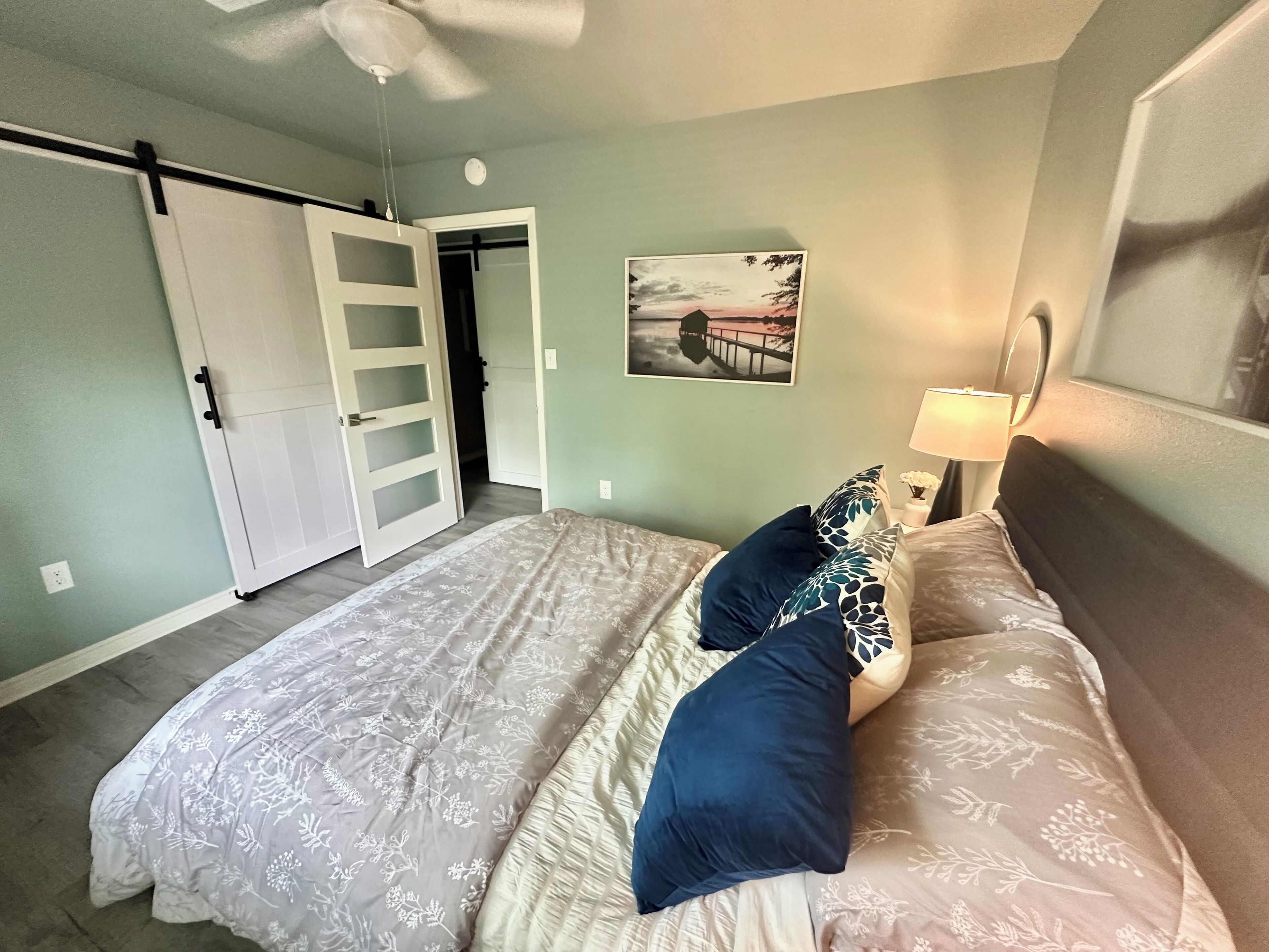 Une chambre à coucher avec un lit, un ventilateur de plafond, une table de chevet avec une lampe et un tableau au mur.