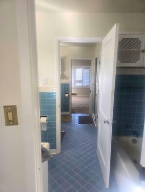 Ein schmaler Korridor mit blau gefliesten Wänden und Boden, der zu einem Badezimmer mit Toilette und Duschbereich führt.