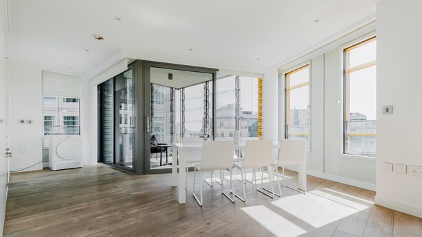 Ein geräumiges Zimmer mit großem Fenster, einem Essbereich mit weißen Stühlen und einer Glastür zum Außenbereich.