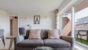 A modern living room with a grey sofa, two cushions, a round glass coffee table, and a view of a brick building through large windows. - Blueground | Bayswater, balcony & w/d, nr parks (London)
