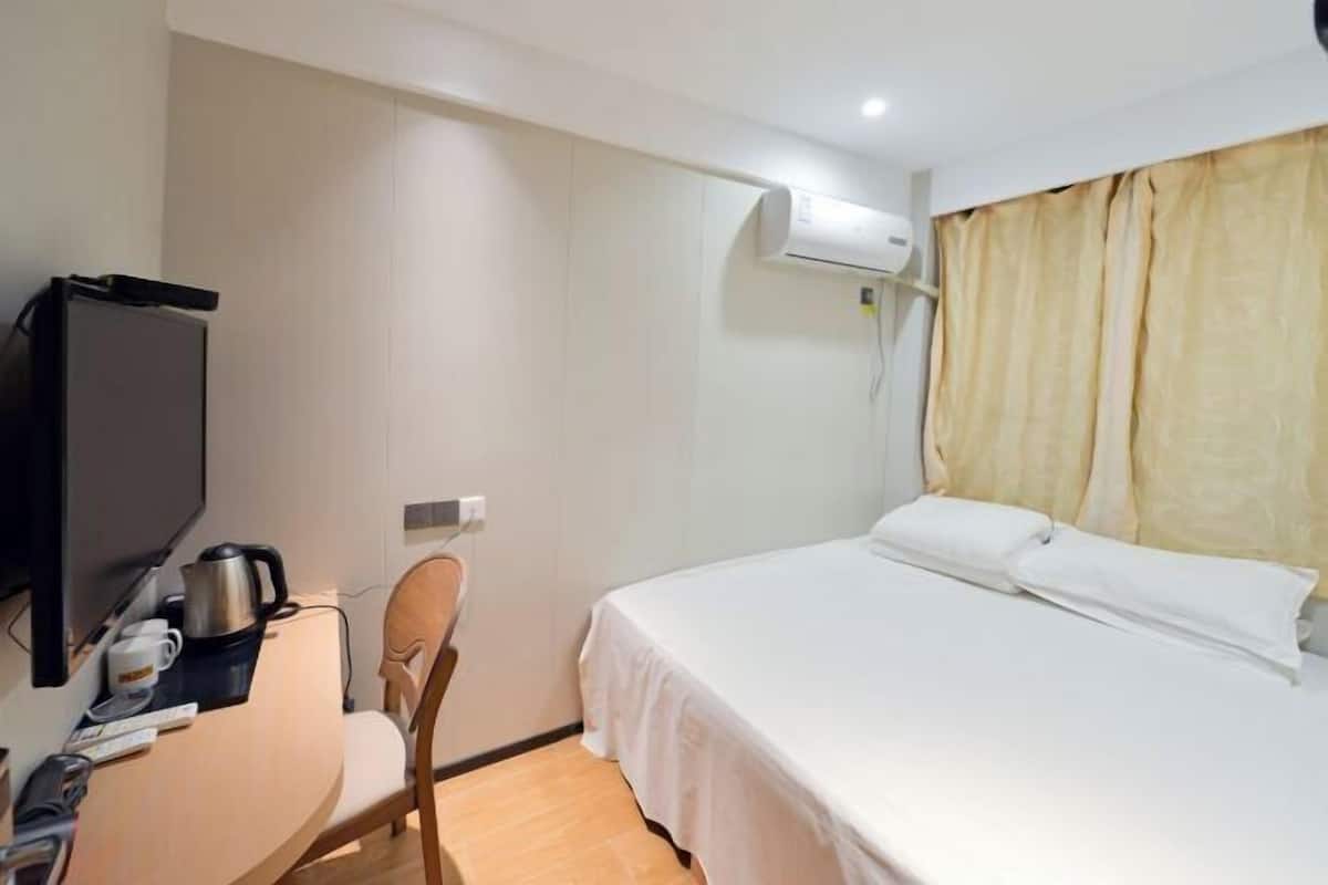 A hotel room with a bed, a desk with a television, a chair, and an air conditioning unit.