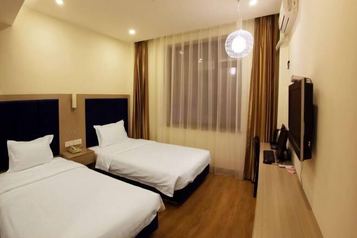 A hotel room with two beds, a wooden floor, a large window with curtains, a flat-screen TV, and a ceiling light fixture.