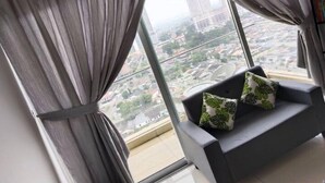 Un salon moderne avec un canapé gris et des coussins à motifs verts, de grandes fenêtres avec des rideaux gris et une vue sur la ville.