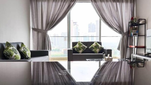Un salon moderne avec une table basse en verre, un canapé gris agrémenté de coussins verts et une vue sur la ville grâce à de grandes fenêtres.