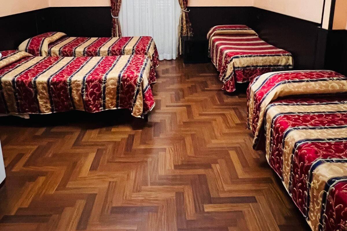 A hotel room with four beds, a wooden herringbone floor, a chandelier, and two paintings on the walls.