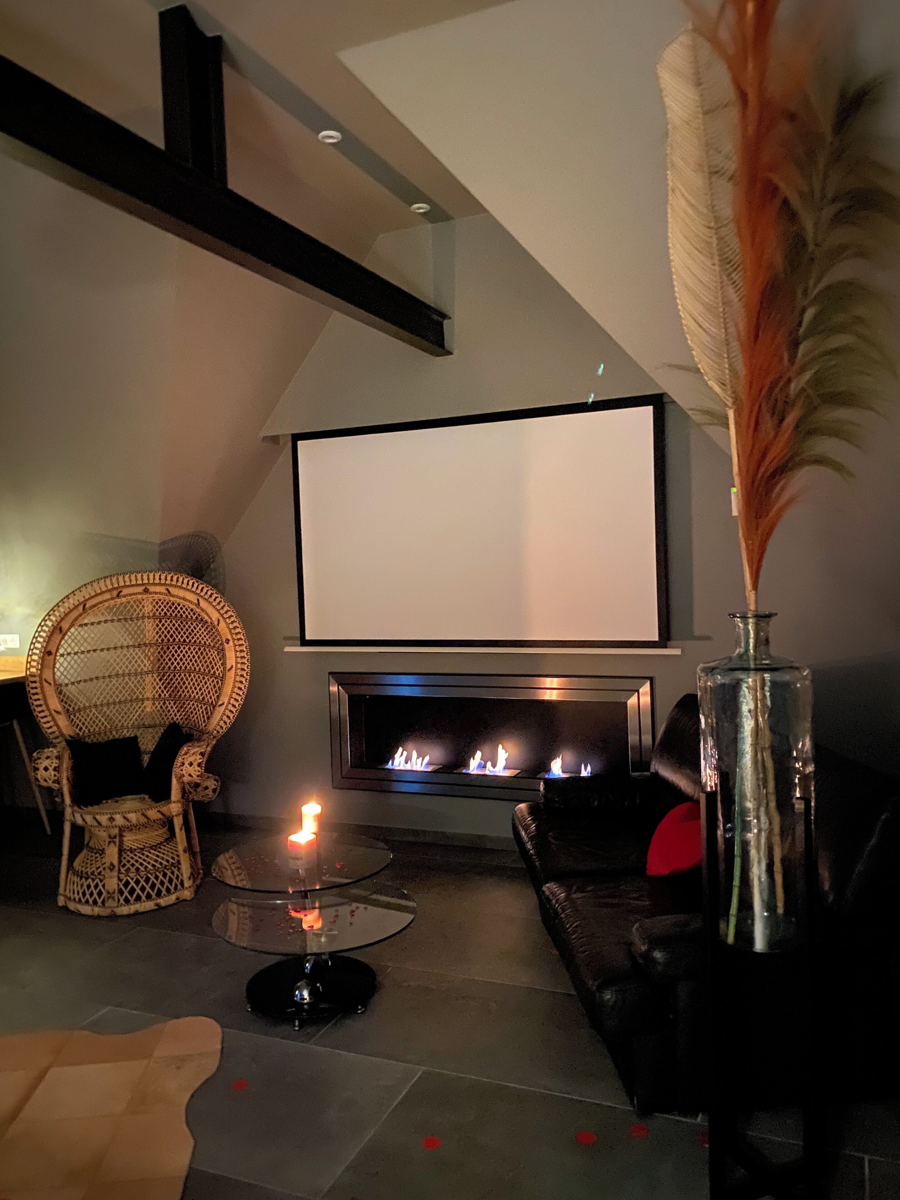 A living room with a fireplace, a television, a wicker chair, a glass table with a candle, and a decorative vase.