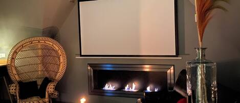 A living room with a fireplace, a television, a wicker chair, a glass table with a candle, and a decorative vase.