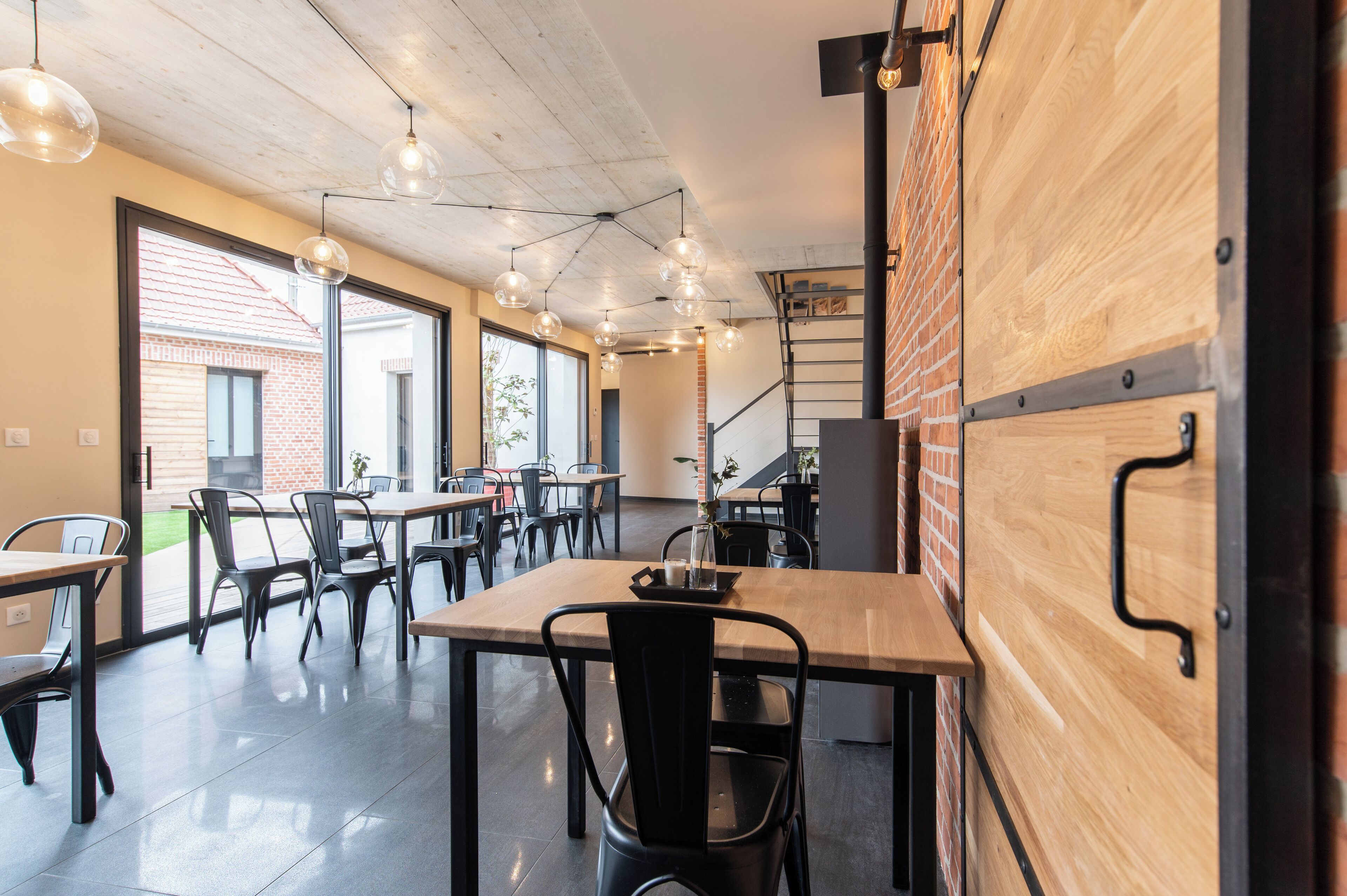 A modern dining area with multiple tables and chairs, large windows, and hanging pendant lights.