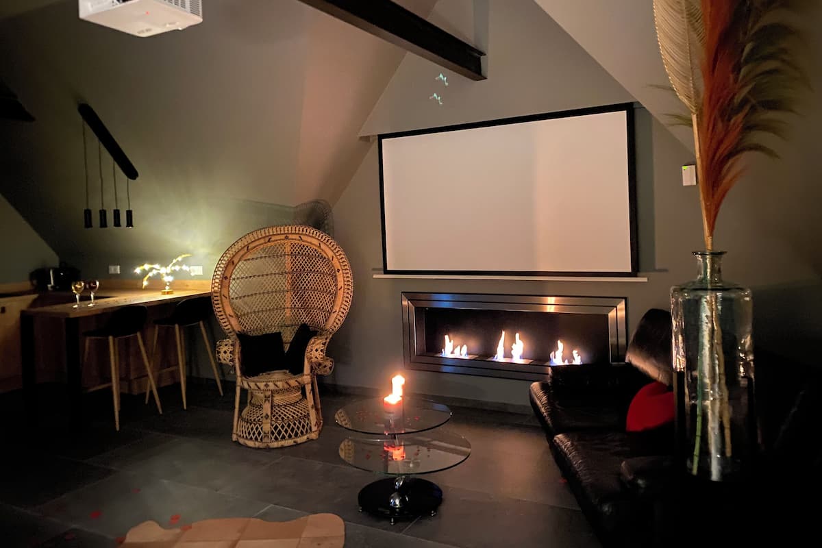 A cozy living room with a fireplace, a large screen, a wicker chair, and a glass coffee table.