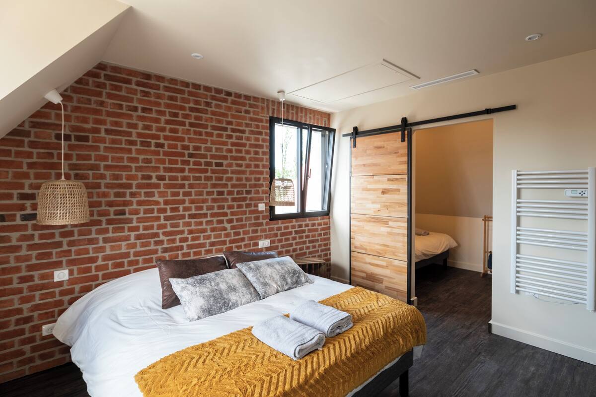 A modern bedroom with a brick wall, a bed with white linens, a yellow throw blanket, and a wooden door.