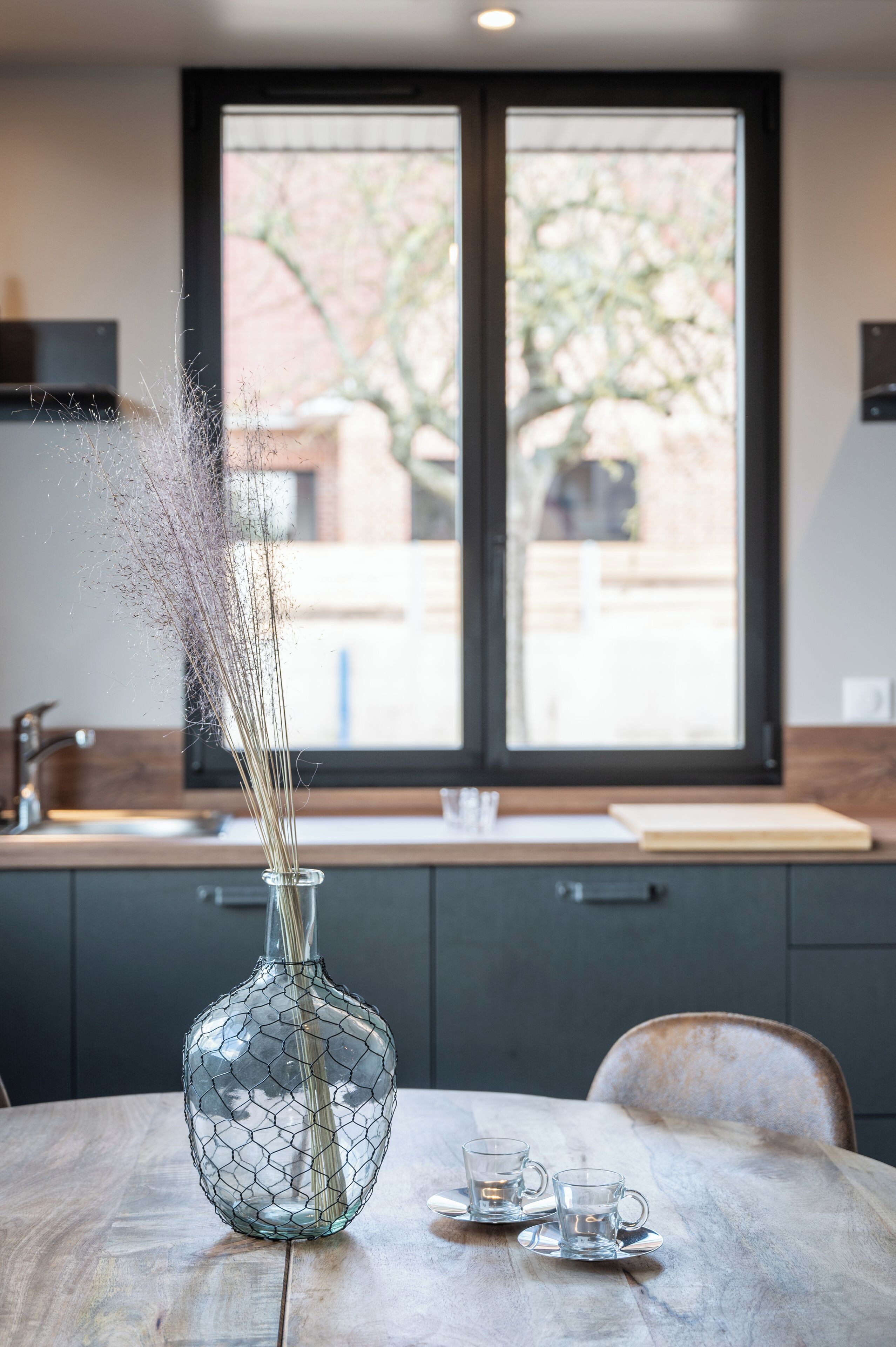 A dining table with a glass vase and two glasses, a window with a view of trees, and a kitchen area in the background.