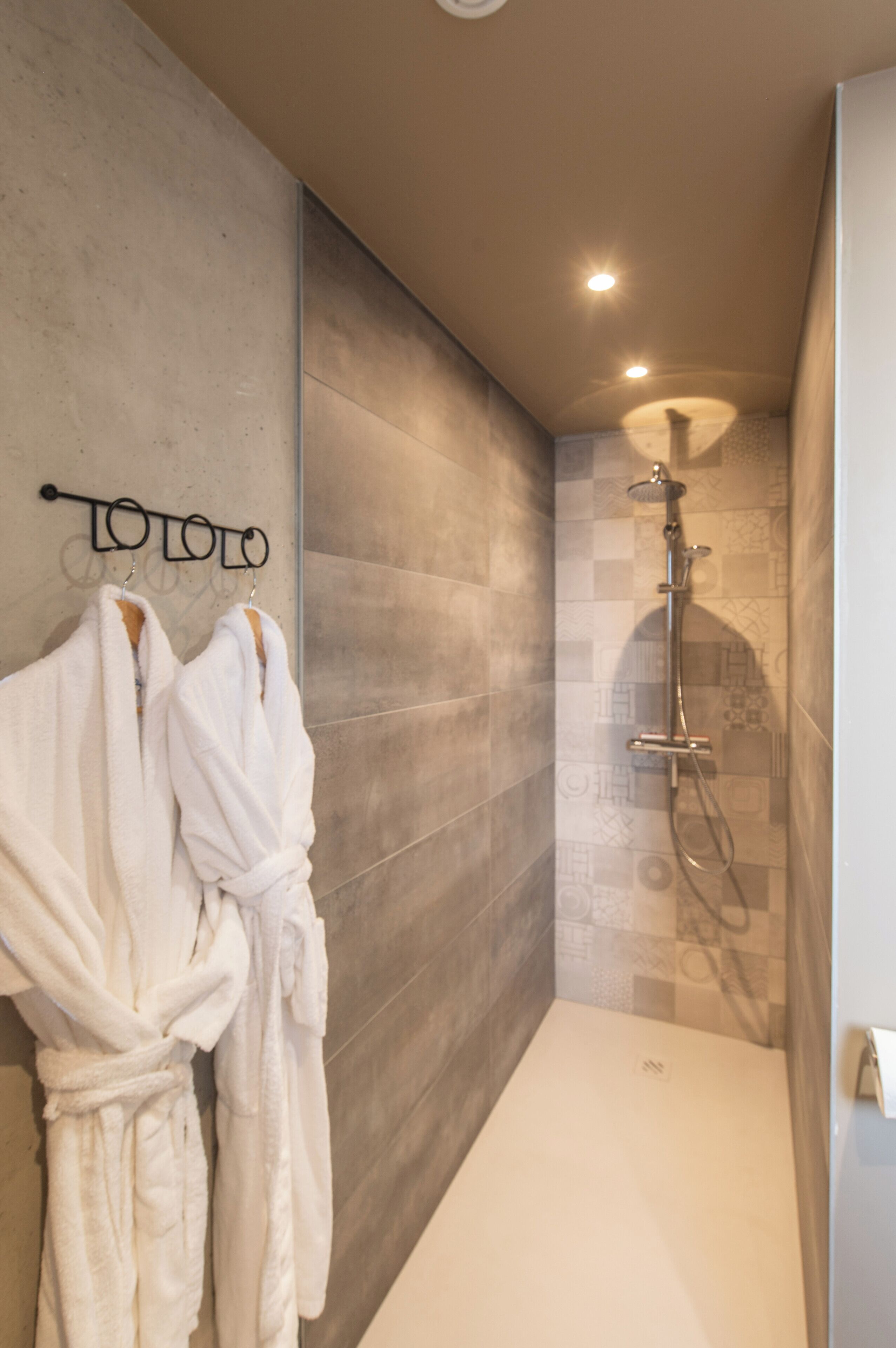 Un baño moderno con ducha, dos albornoces blancos colgados en un perchero y un diseño de pared texturizado.