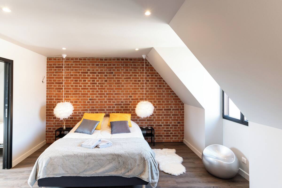 A modern bedroom with a brick wall, a bed with yellow and gray pillows, a fluffy white chair, and a round white stool.