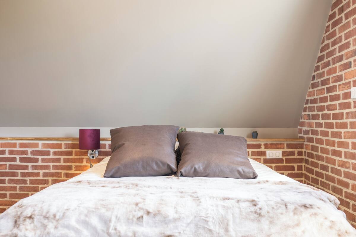 A bedroom with a brick wall, a bed with two pillows, a small plant, and a purple lamp.