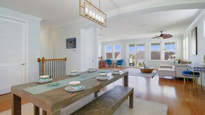 A spacious dining area with a long wooden table set for a meal, a ceiling fan, and a view of the outdoors through large windows. - Villa Vilano |Summer by the beach! (St. Augustine Vilano Beach)