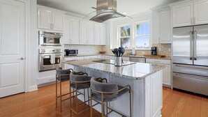 A modern kitchen with white cabinetry, stainless steel appliances, a central island with bar stools, and a view of the outdoors through large windows. - Villa Vilano |Summer by the beach! (St. Augustine Vilano Beach)