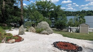 A sandy outdoor area with large rocks and mulch, surrounded by trees and a lake view. - Charming Grand View/Lake Loft in Brewster with WiFi, AC (Brewster)