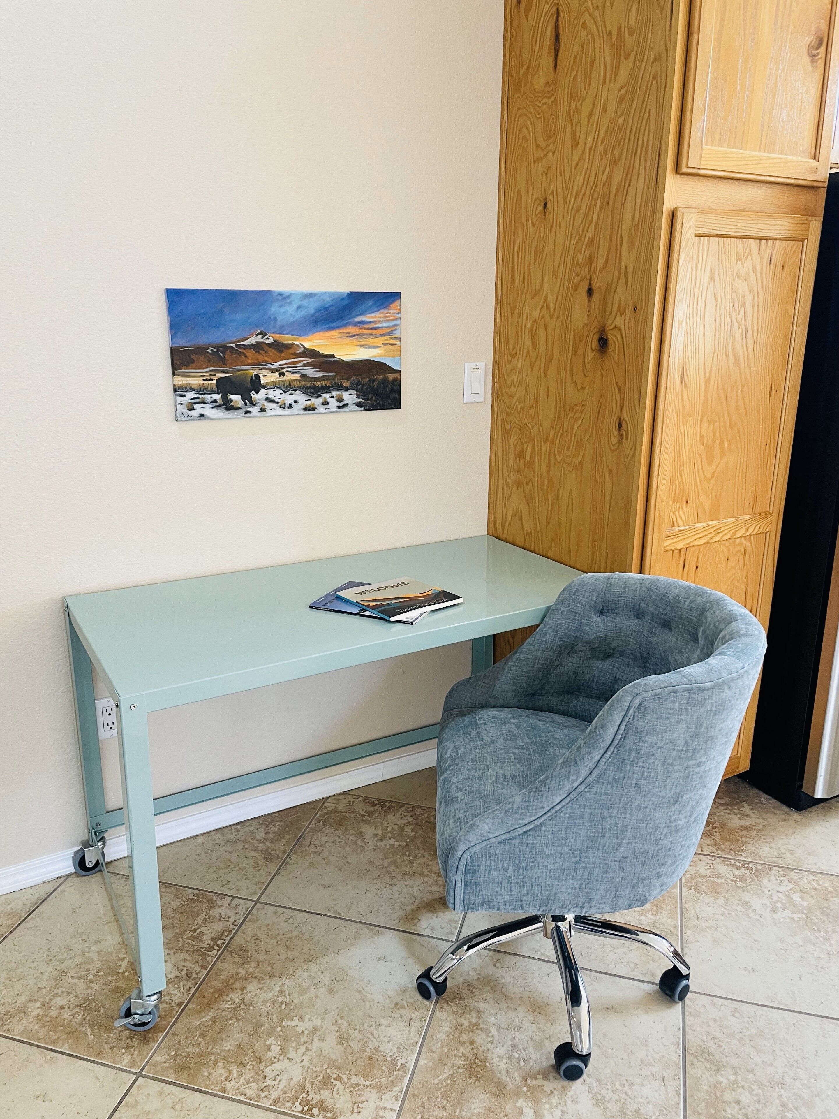 A room with a light blue desk, a grey office chair, a painting on the wall, and a wooden wardrobe.