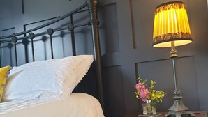 A bed with a white bedspread and pillows, a nightstand with a lamp and a vase of flowers, and a dark wall with geometric paneling. - The Cube hidden gem in the heart of ilfracombe , 2 minute walk to tunnels beach! (Ilfracombe)
