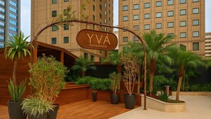Μια πινακίδα εστιατορίου YVA, περιτριγυρισμένη από φυτά σε γλάστρες και φοίνικες, με ένα σύγχρονο κτίριο στο βάθος.