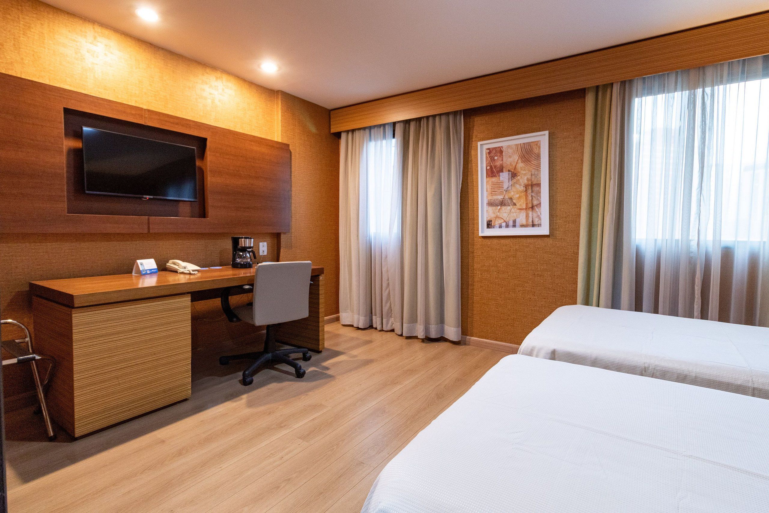 A hotel room with a wooden desk, a flat-screen TV, a bed, and a window with curtains.