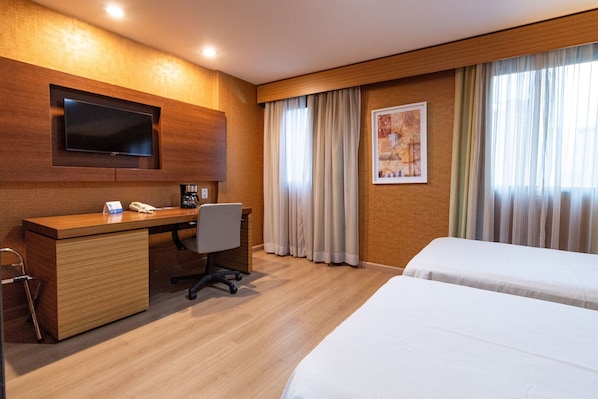 Quarto de hotel com escrivaninha de madeira, televisão de tela plana, cama e janela com cortinas.