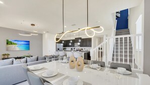 A modern dining area with a white table set for six, a gray sofa, and a kitchen in the background.