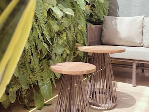 Two round wooden tables with woven bases, a sofa with a cushion, and lush green foliage in the background. - Setai Yacht - Garden III - By Almare (João Pessoa)