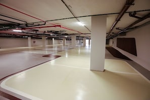 An empty parking garage with white floors, red and black pipes on the ceiling, and white pillars. - Setai Yacht - Garden III - By Almare (João Pessoa)