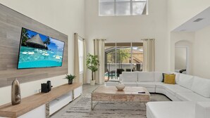 Ein geräumiges Wohnzimmer mit einem großen Flachbildfernseher, einem weißen Modulsessel, einem Holztisch und einem großen Fenster mit Vorhängen.