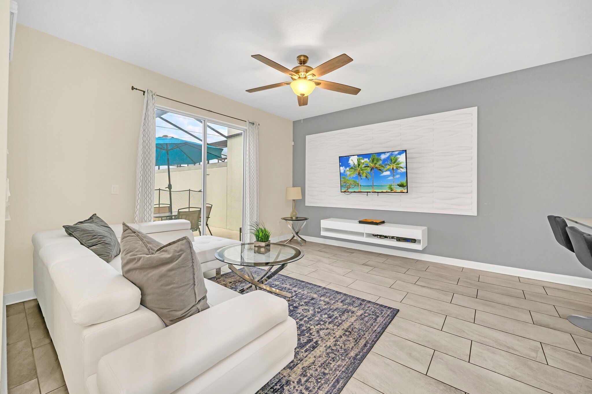 A modern living room with a white sofa, a glass coffee table, a ceiling fan, and a TV mounted on a white wall.