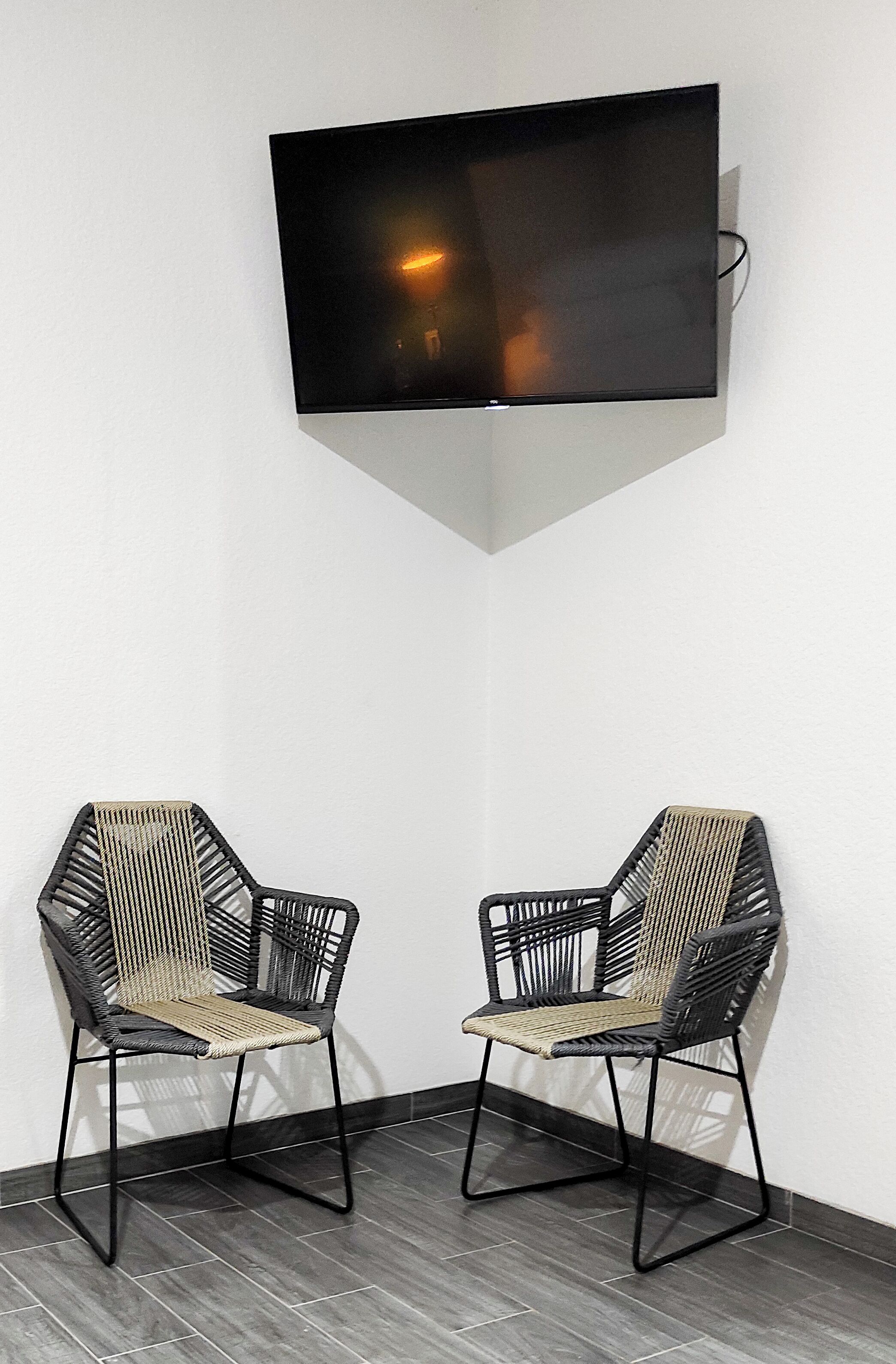 Ein Eckzimmer mit einem an der Wand befestigten Flachbildfernseher, zwei schwarze Gestellstühle mit gewobenen Sitzflächen und dunkelgrauem Fliesenboden.